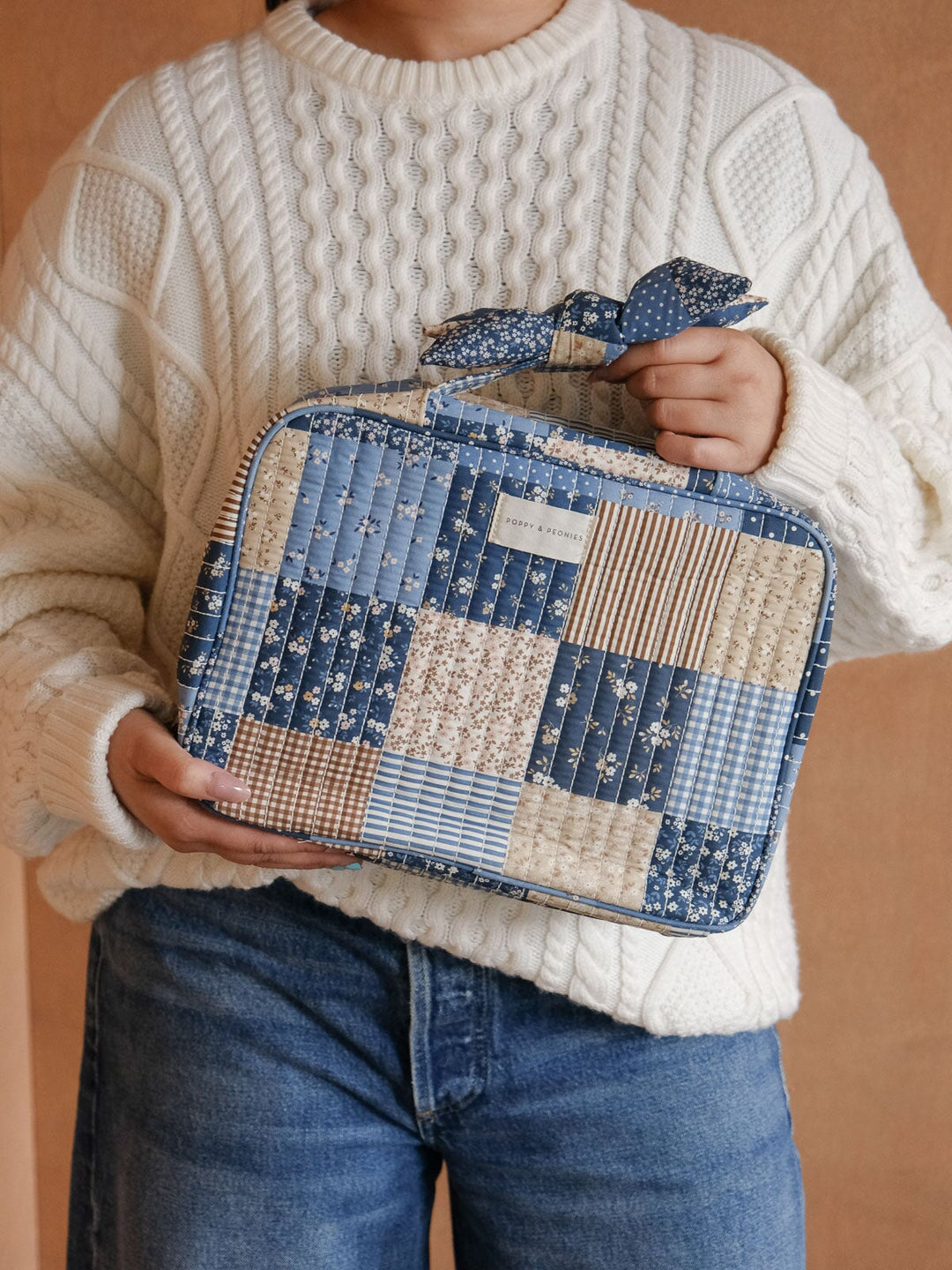 Lifestyle close-up of the Hook Me Up Cosmetic Case in Navy Patchwork, highlighting its mix of blue and neutral patchwork prints, zip-around closure, and signature bow handle.