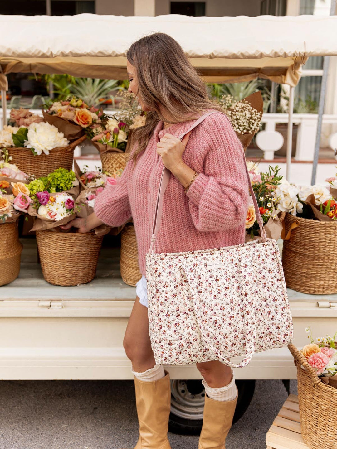 Woman in pink sweater and tan boots holding a floral-patterned tote bag in front of a flower truck.