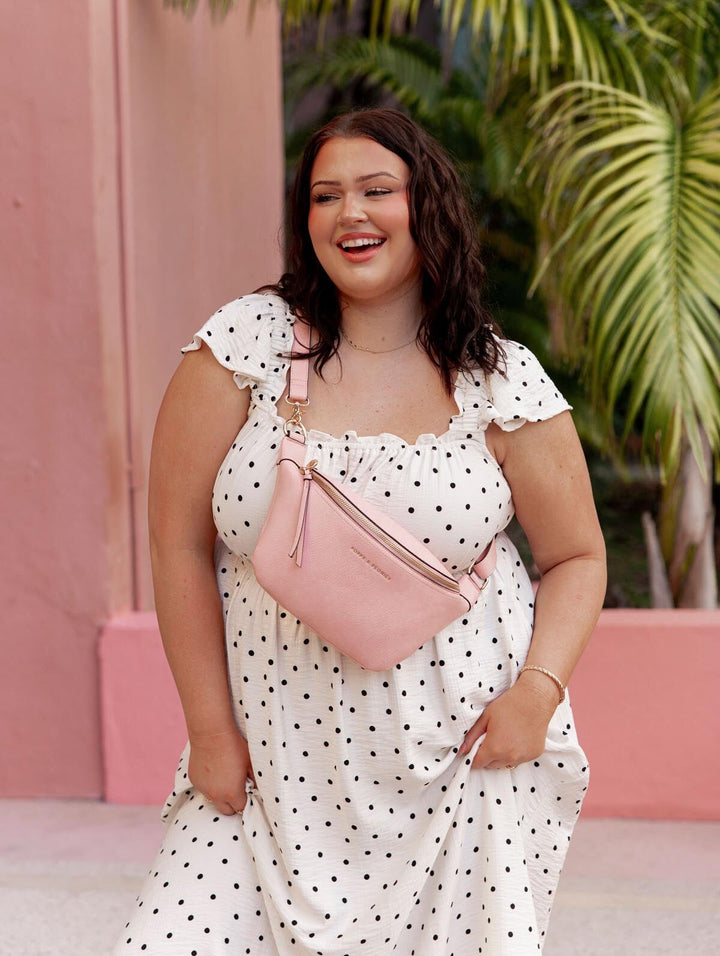Woman in a polka dot dress with a pink bag standing in front of a pink wall with greenery.
