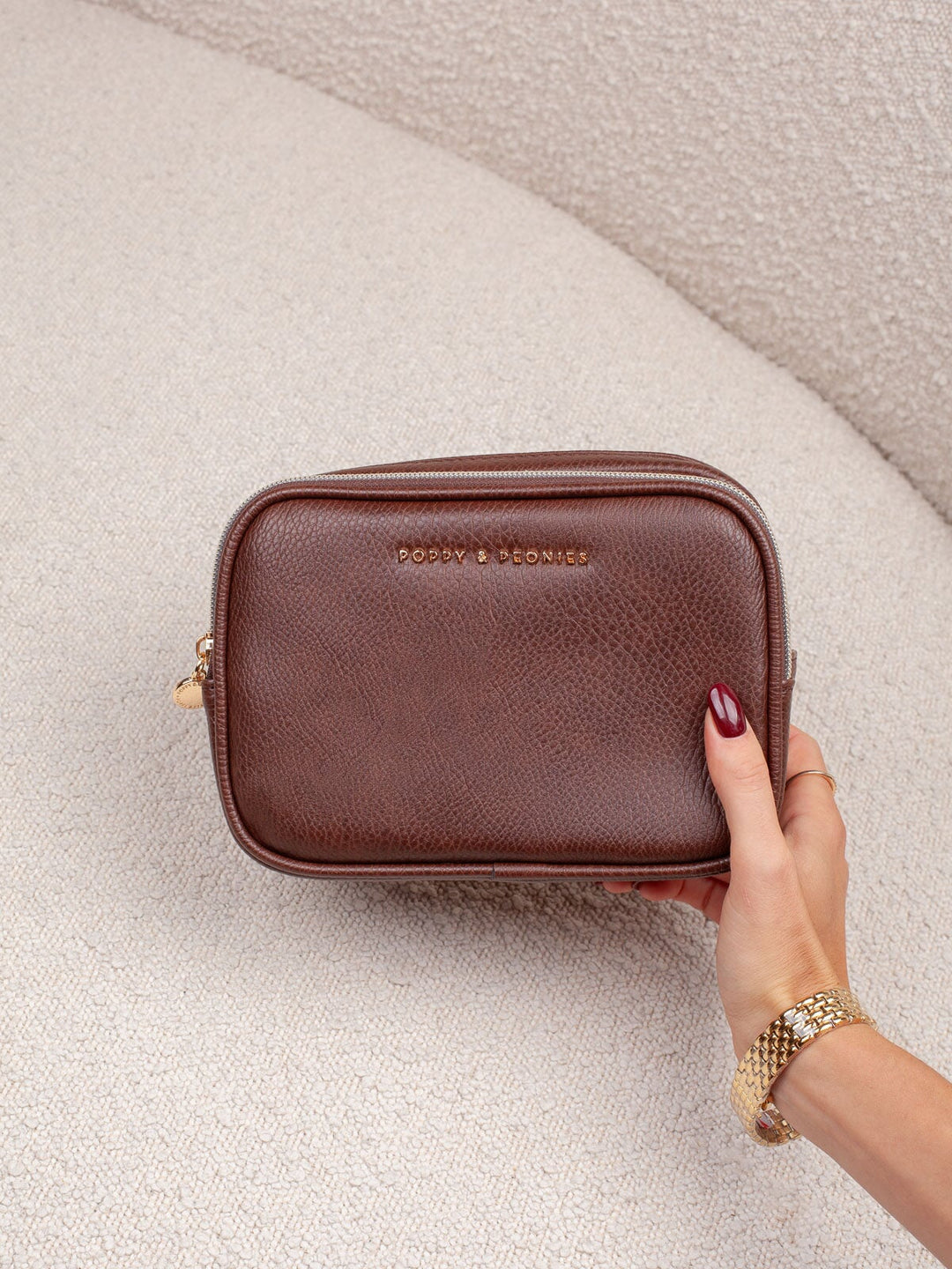 Handheld lifestyle shot of the Poppy & Peonies Pretty Connected Case in Espresso, showcasing its rich espresso exterior, compact structured silhouette, double-zip design, and gold-tone branding.