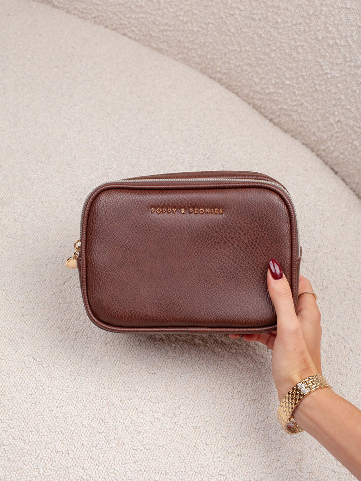 Handheld lifestyle shot of the Poppy & Peonies Pretty Connected Case in Espresso, showcasing its rich espresso exterior, compact structured silhouette, double-zip design, and gold-tone branding.