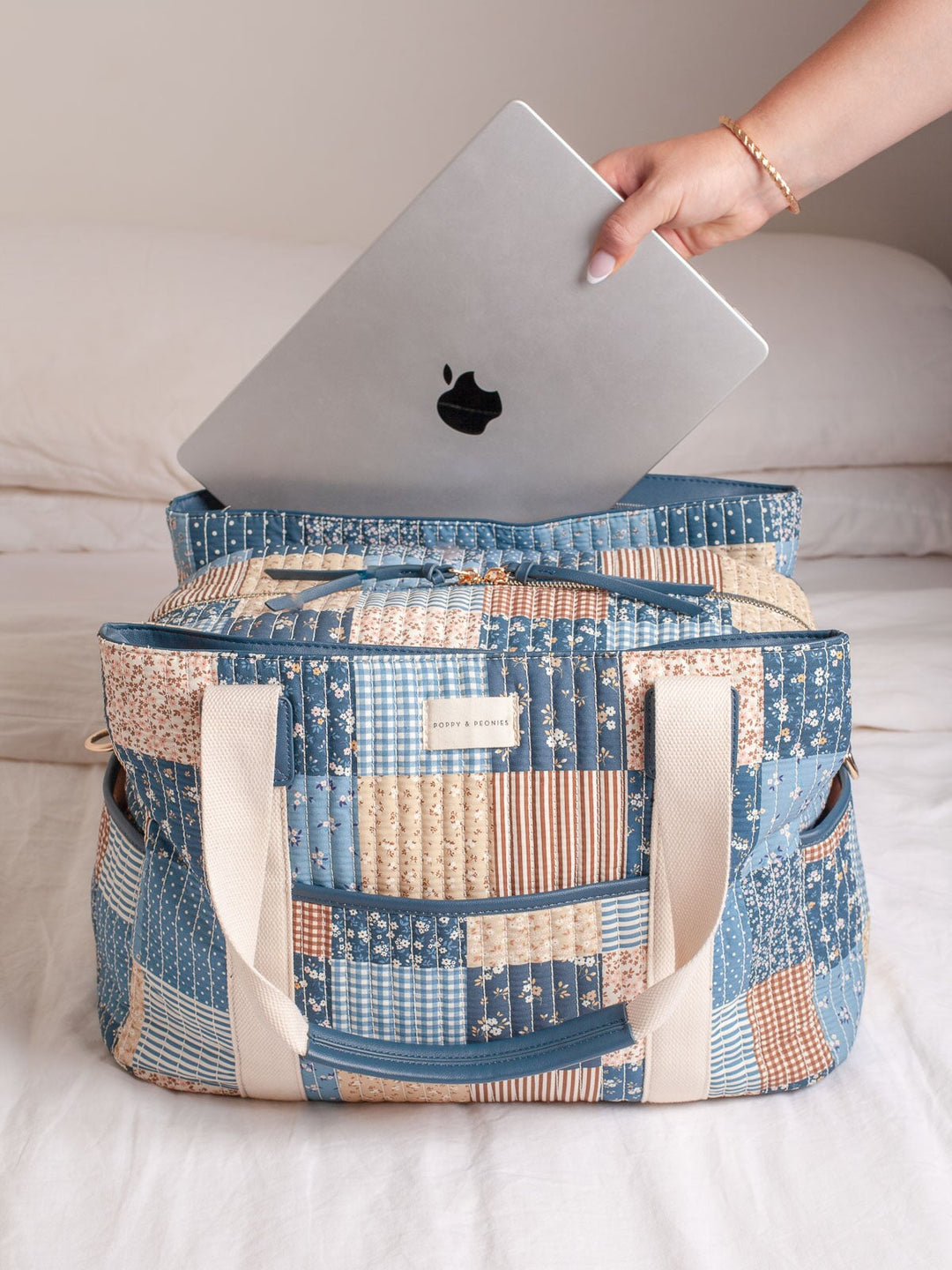 Lifestyle shot of the Poppy & Peonies Mini Voyager in Navy Patchwork, shown with a laptop being placed inside to highlight its spacious interior, laptop-friendly storage, quilted patchwork exterior, front pocket, and sturdy shoulder straps.