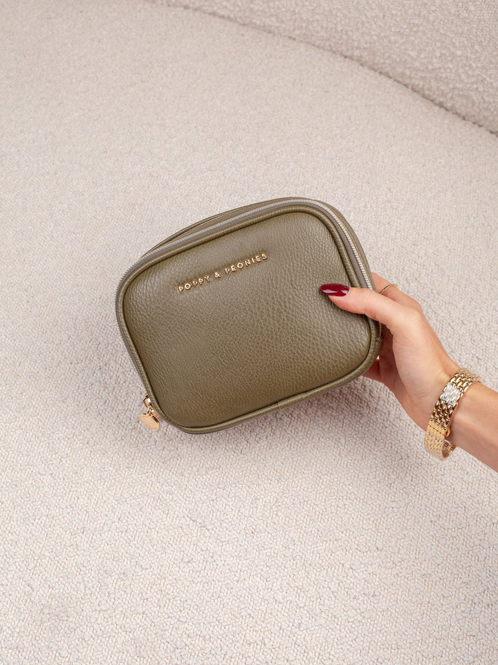 Handheld lifestyle shot of the Poppy & Peonies Pretty Connected Case in Olive, showcasing its rich olive exterior, compact structured silhouette, double-zip design, and gold-tone branding.