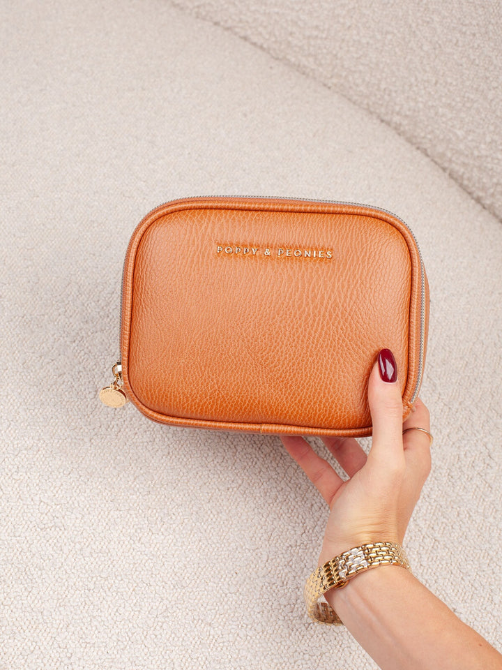 Handheld lifestyle shot of the Poppy & Peonies Pretty Connected Case in Cognac, showcasing its rich cognac exterior, compact structured silhouette, double-zip design, and gold-tone branding.