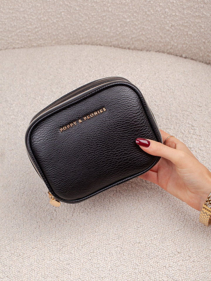 Handheld lifestyle shot of the Poppy & Peonies Pretty Connected Case in Black, showcasing its rich navy exterior, compact structured silhouette, double-zip design, and gold-tone branding.