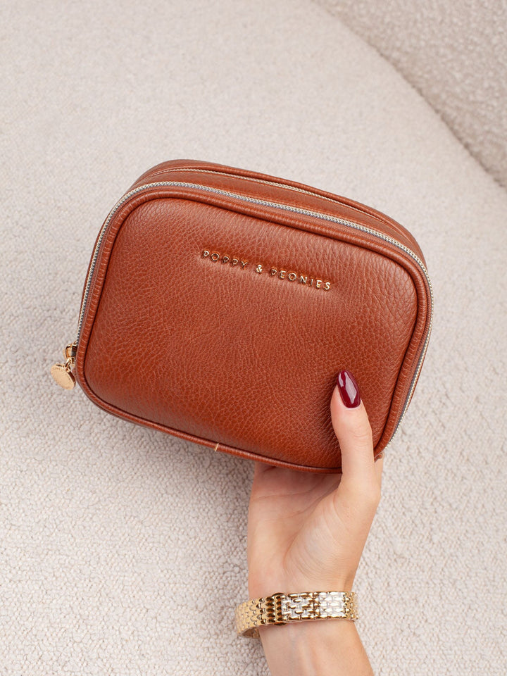 Handheld lifestyle shot of the Poppy & Peonies Pretty Connected Case in Chestnut, showcasing its rich chestnut exterior, compact structured silhouette, double-zip design, and gold-tone branding.