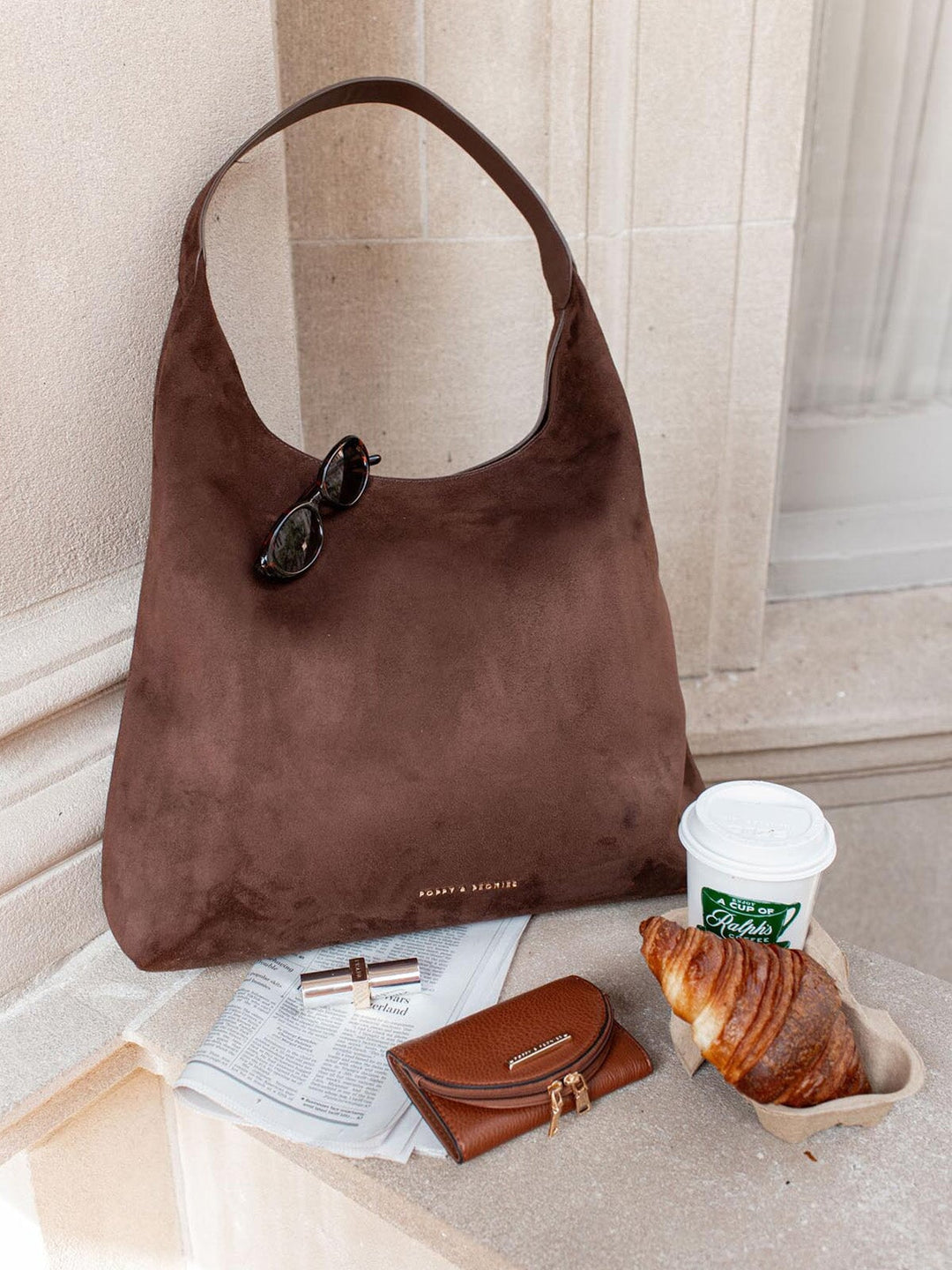 Brown vegan suede hobo style handbag with sunglasses, coffee cup, and croissant on a marble surface.