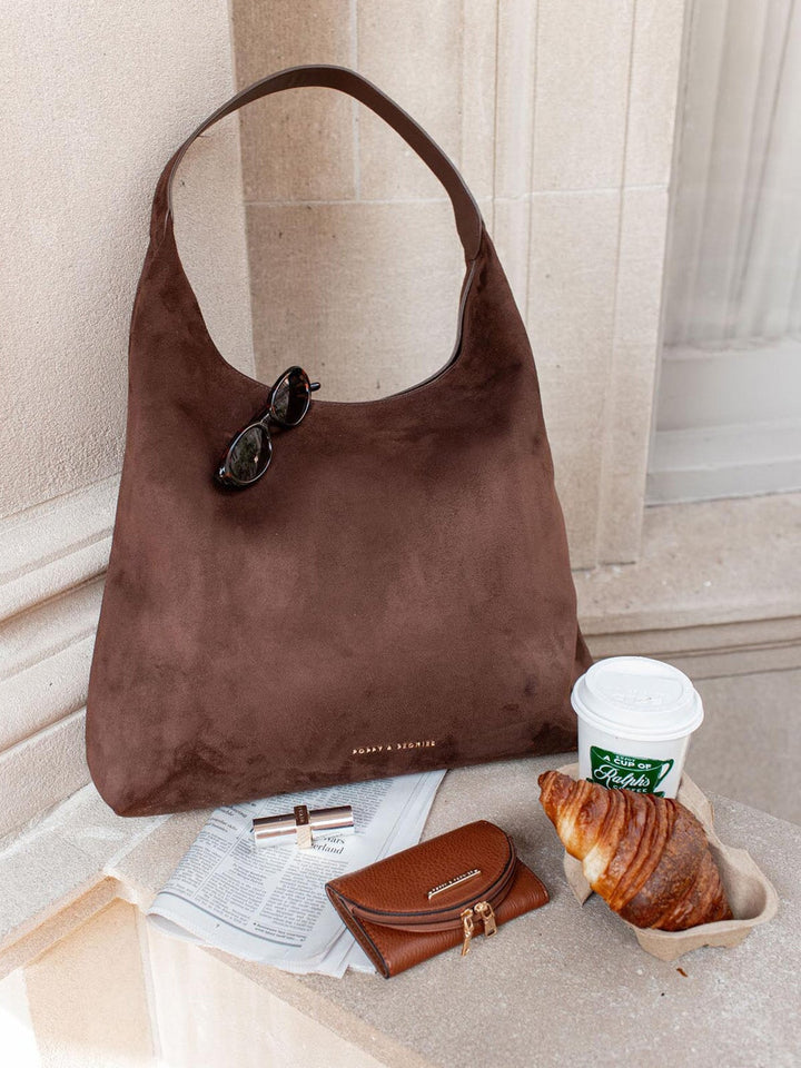 Brown vegan suede hobo style handbag with sunglasses, coffee cup, and croissant on a marble surface.