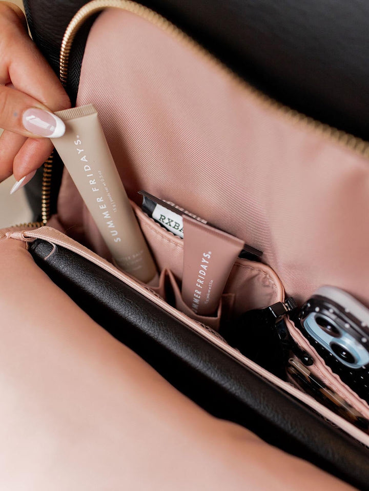 Hand holding a skincare product over an open backpack pocket containing other items.