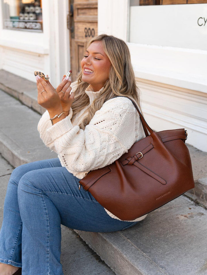 Poppy & Peonies The Knot Tote in Chestnut, worn over the shoulder by a woman in a cream knit sweater and blue jeans while sitting on storefront steps and applying lipstick.