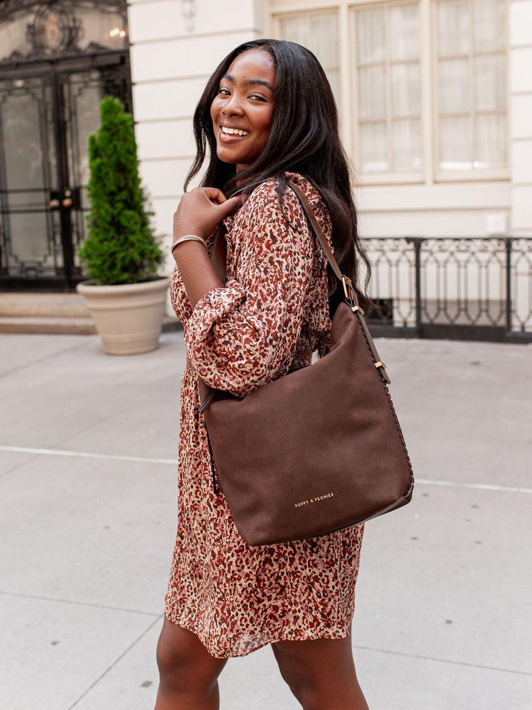 Woman in a patterned dress holding a vegan suede brown handbag outdoors.