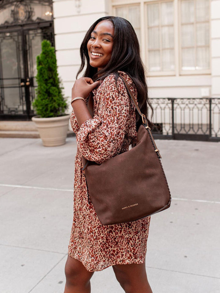 Woman in a patterned dress holding a vegan suede brown handbag outdoors.