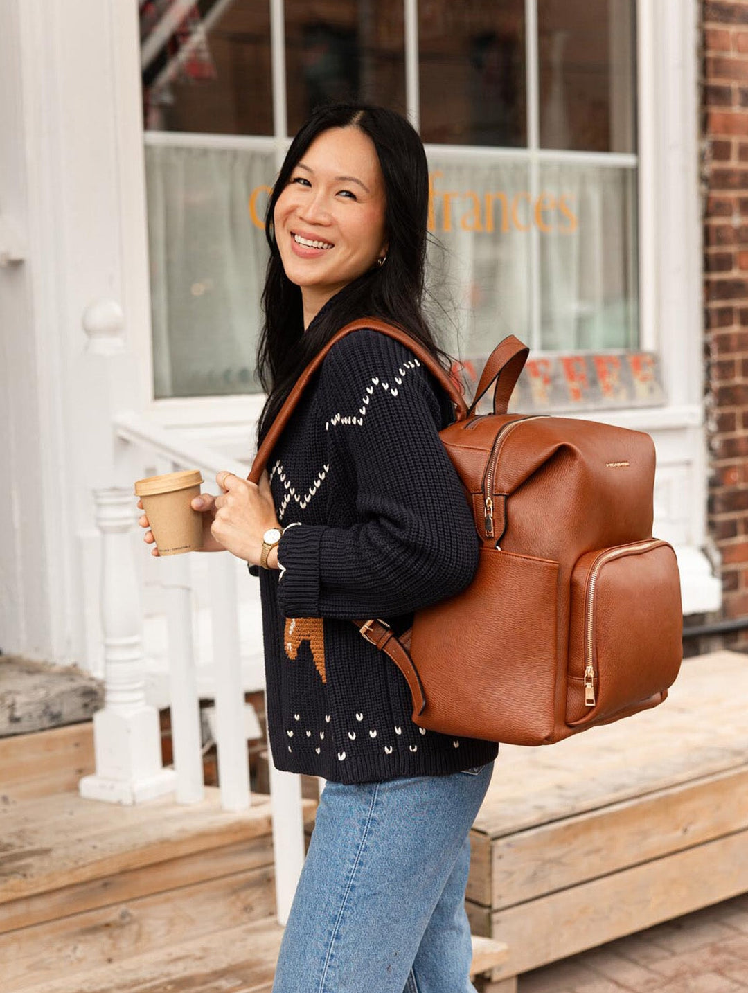 Poppy & Peonies The Cabin Backpack in Chestnut worn over both shoulders by a smiling woman holding a takeaway coffee outside a café.