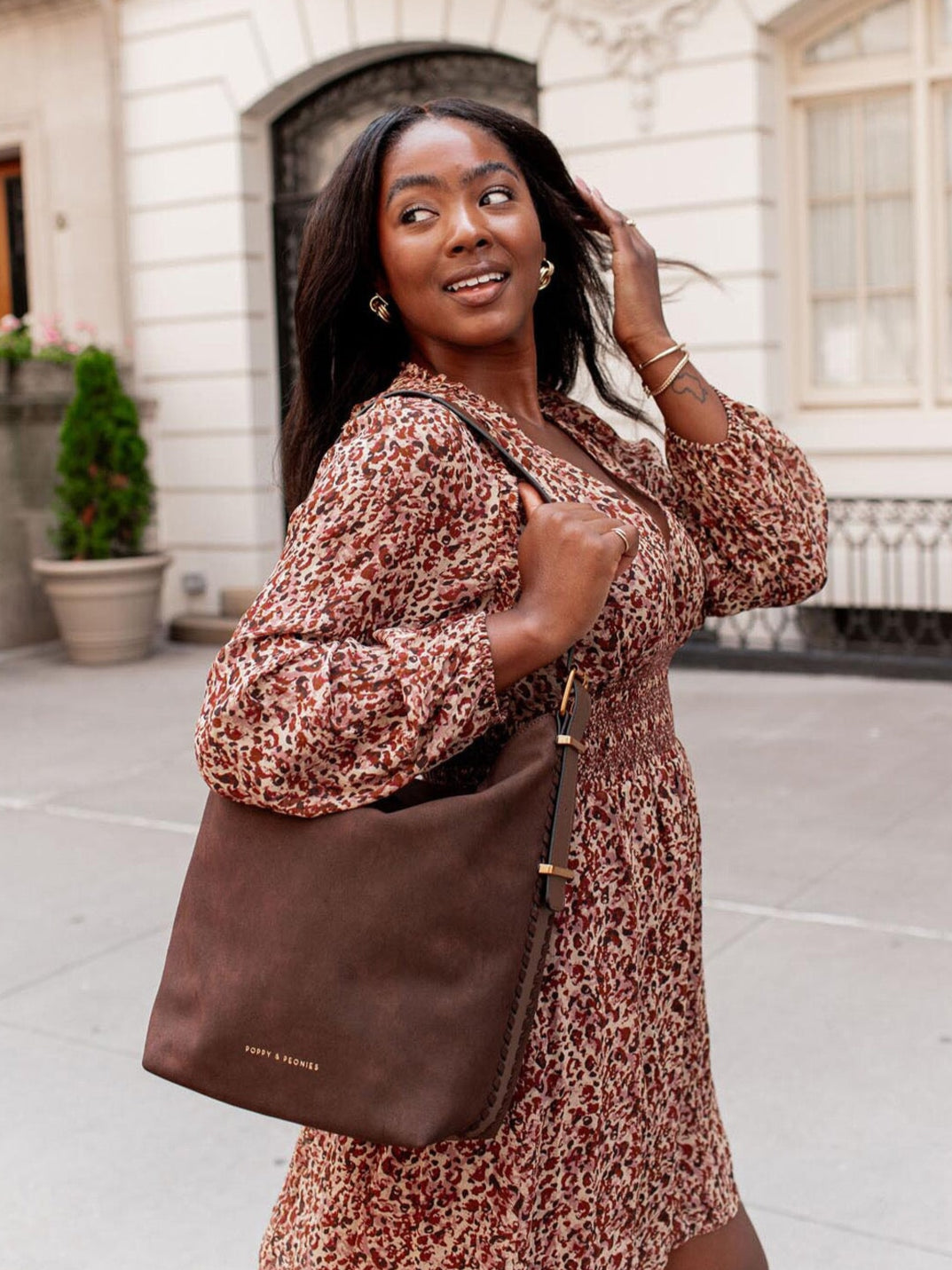Woman in a printed dress holding a brown vegan suede bag outdoors.