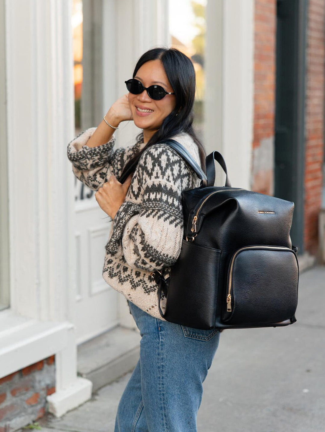 Poppy & Peonies The Cabin Backpack in Black worn on the back by a smiling woman in sunglasses and a patterned sweater outside a storefront.