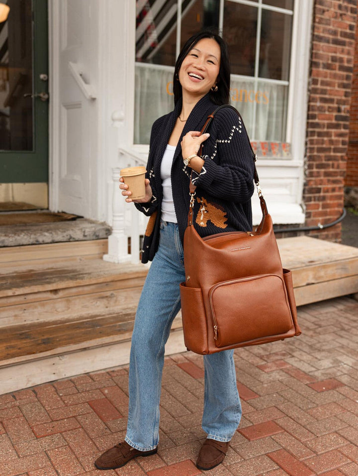 Poppy & Peonies The Cabin Backpack in Chestnut styled with a navy cardigan and jeans as a smiling woman carries it by the top handle on a brick sidewalk.