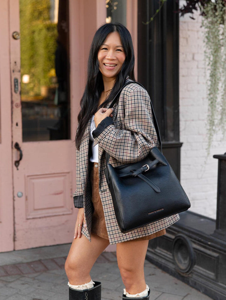 Poppy & Peonies The Knot Shoulder Bag in Black worn over the shoulder by a smiling woman in a plaid blazer, highlighting the bag's relaxed shape and gold-tone hardware.