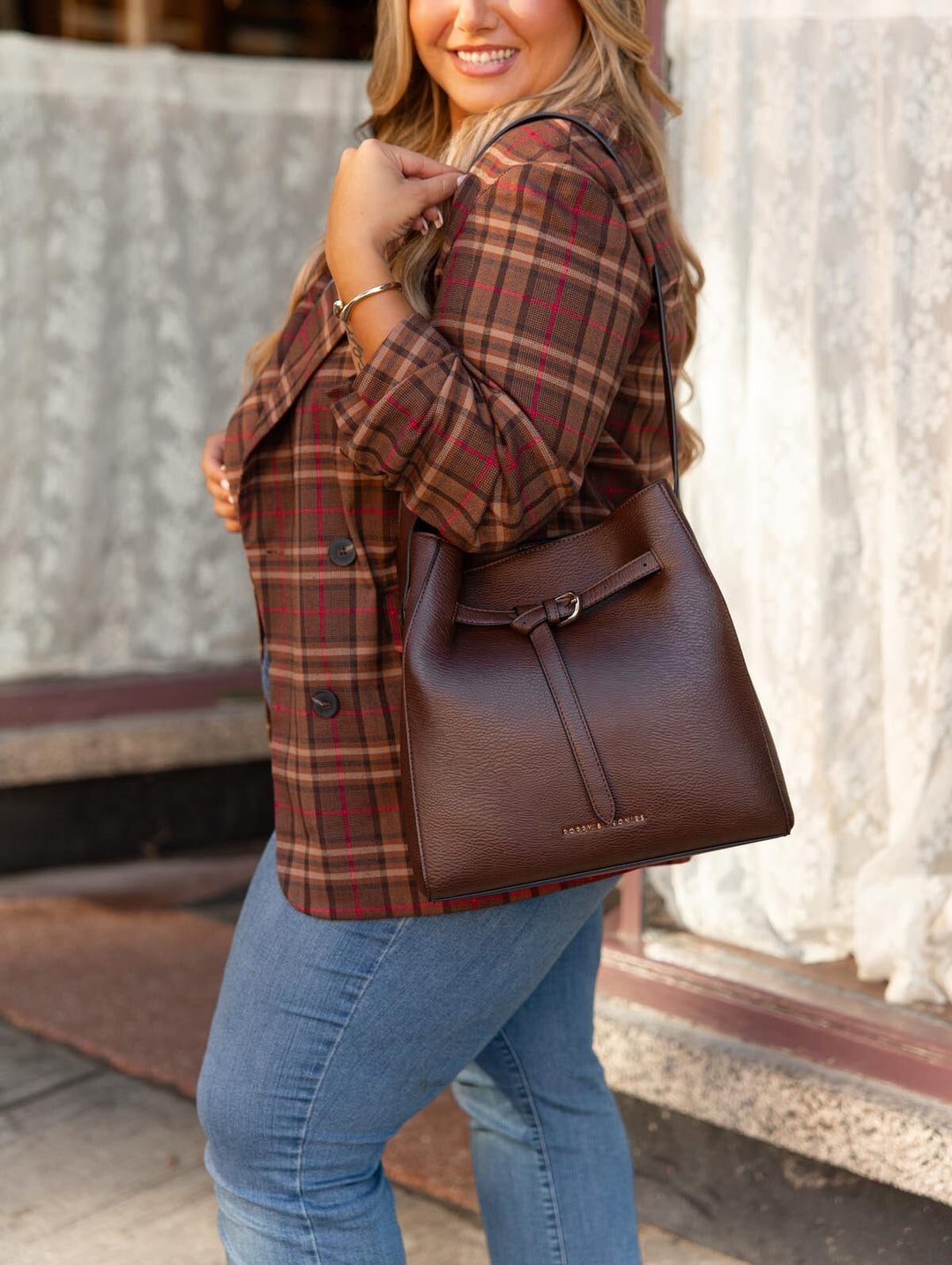 Poppy & Peonies The Knot Shoulder Bag in Espresso worn over the shoulder by a woman in a brown plaid blazer and blue jeans outside a storefront.