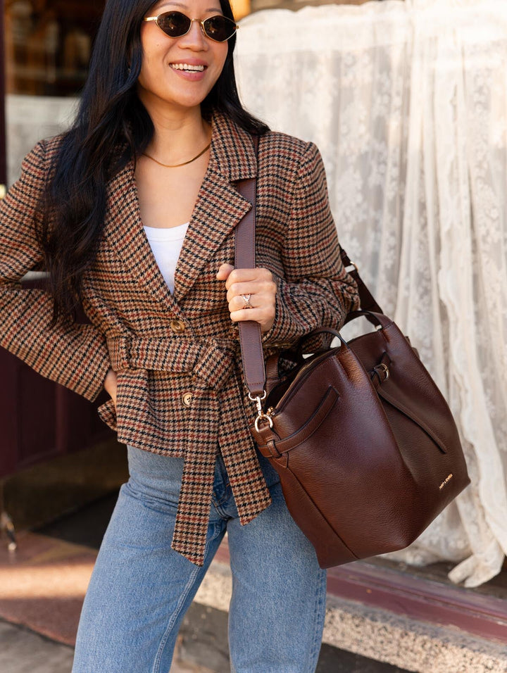 Poppy & Peonies The Knot Tote in Espresso, carried over the shoulder by a woman wearing a brown plaid blazer and white top, highlighting its pebbled texture and long handles.