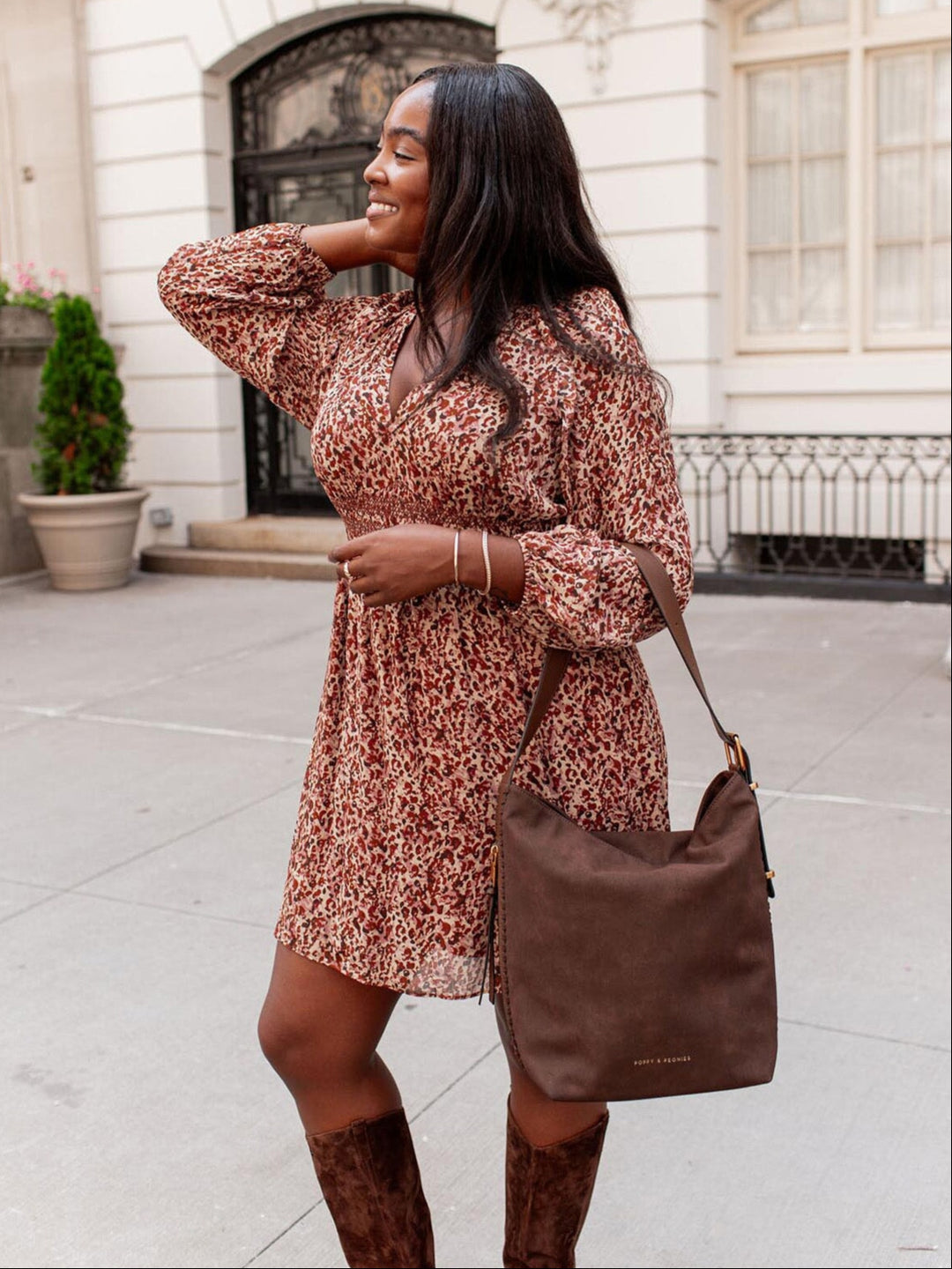 Woman in a patterned dress holding a vegan suede brown bag on a city street.