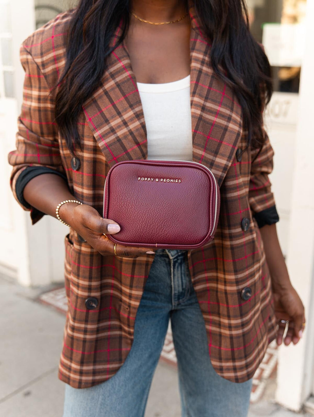 Close-up of the Poppy & Peonies Pretty Connected Case in Berry held in front of a plaid blazer, showcasing its compact shape and zip-around design.