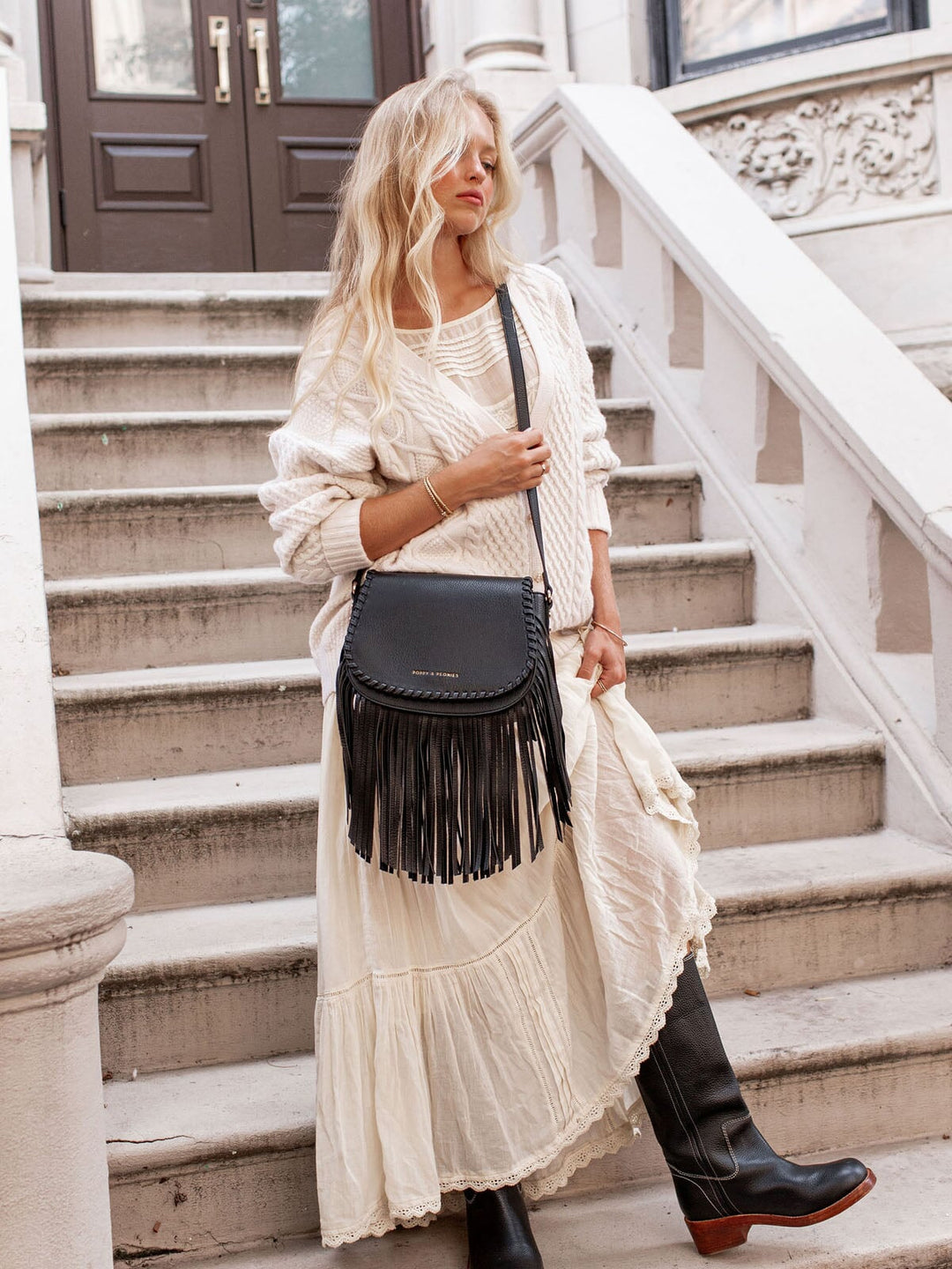 Woman in a white outfit with a black fringed bag standing on steps.