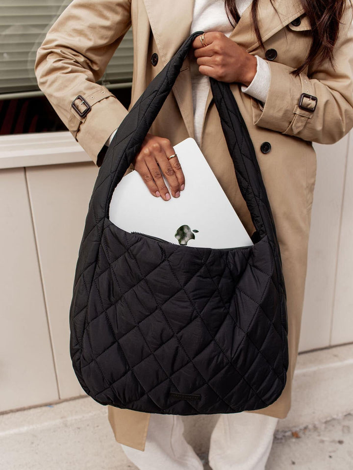 Person holding a black quilted handbag with a beige coat and white pants.