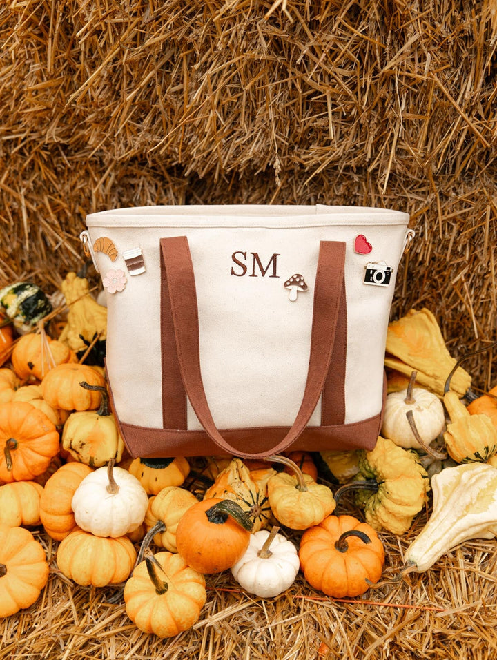 Poppy & Peonies letter patches and decorative pins displayed on the Signature tote in Chestnut, styled among pumpkins and hay bales at a pumpkin patch.