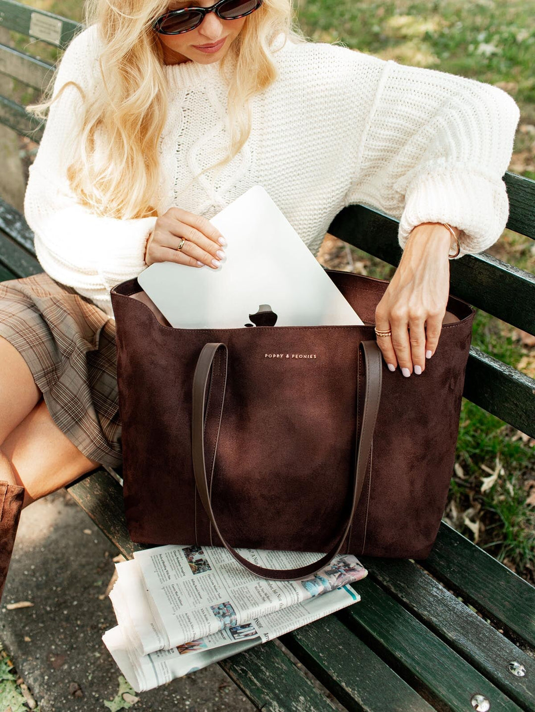 Woman sitting on a bench with a brown leather tote bag, wearing sunglasses and a white sweater.