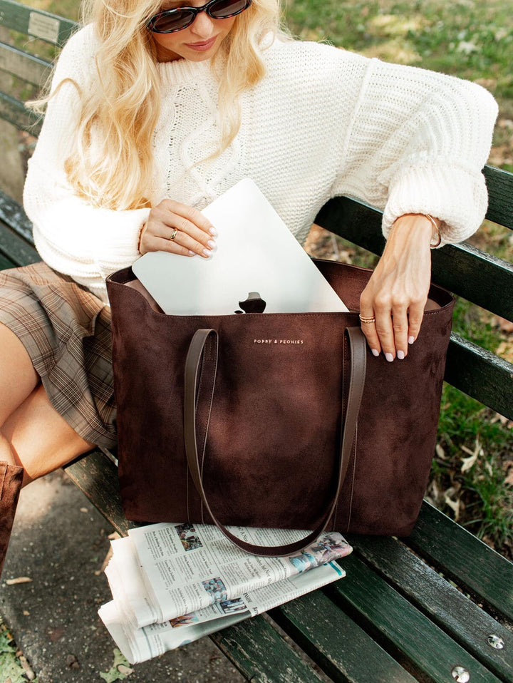 Woman sitting on a bench with a brown leather tote bag, wearing sunglasses and a white sweater.