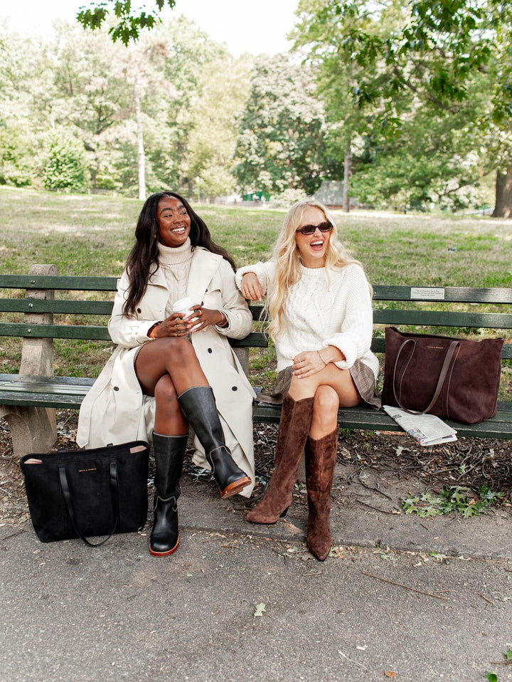 Two women sitting on a park bench with vegan leather tote bags and trees and grass in the background