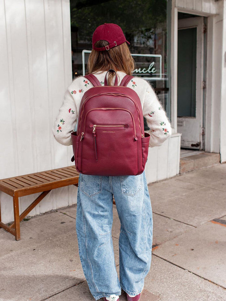 Person wearing a berry coloured vegan leather backpack and red cap walking on a sidewalk.
