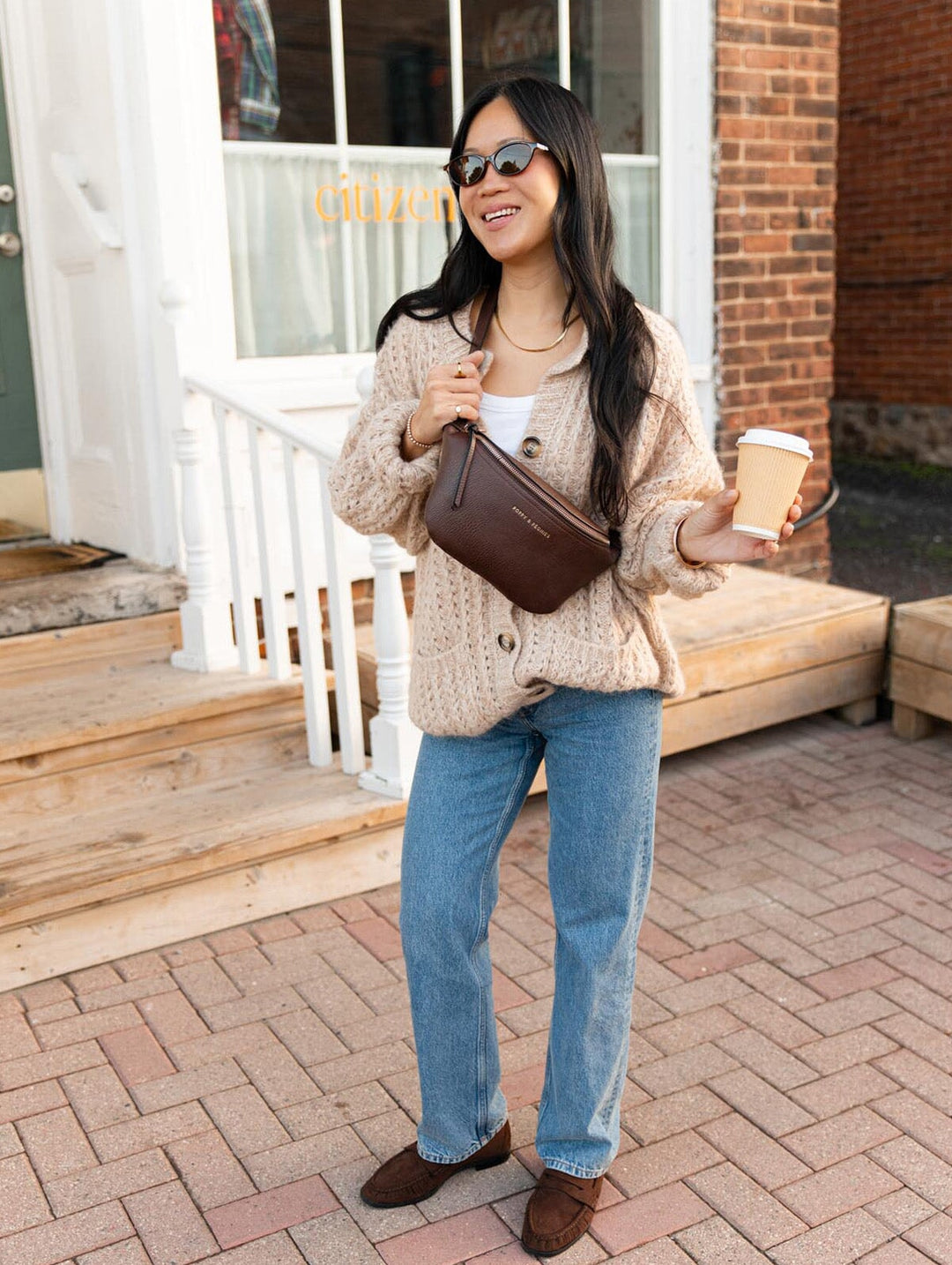 Poppy & Peonies The Sidekick in Espresso styled across the chest, featuring a rich brown exterior, front zipper pocket, and adjustable strap in a casual coffee shop setting.