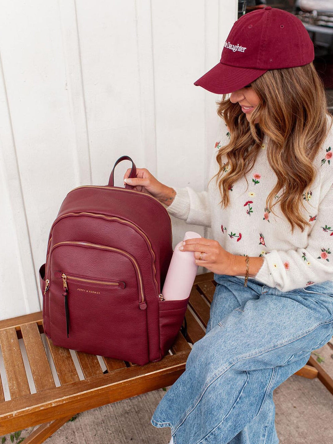 Woman sitting on a wooden bench with a berry coloured backpack and pink water bottle.