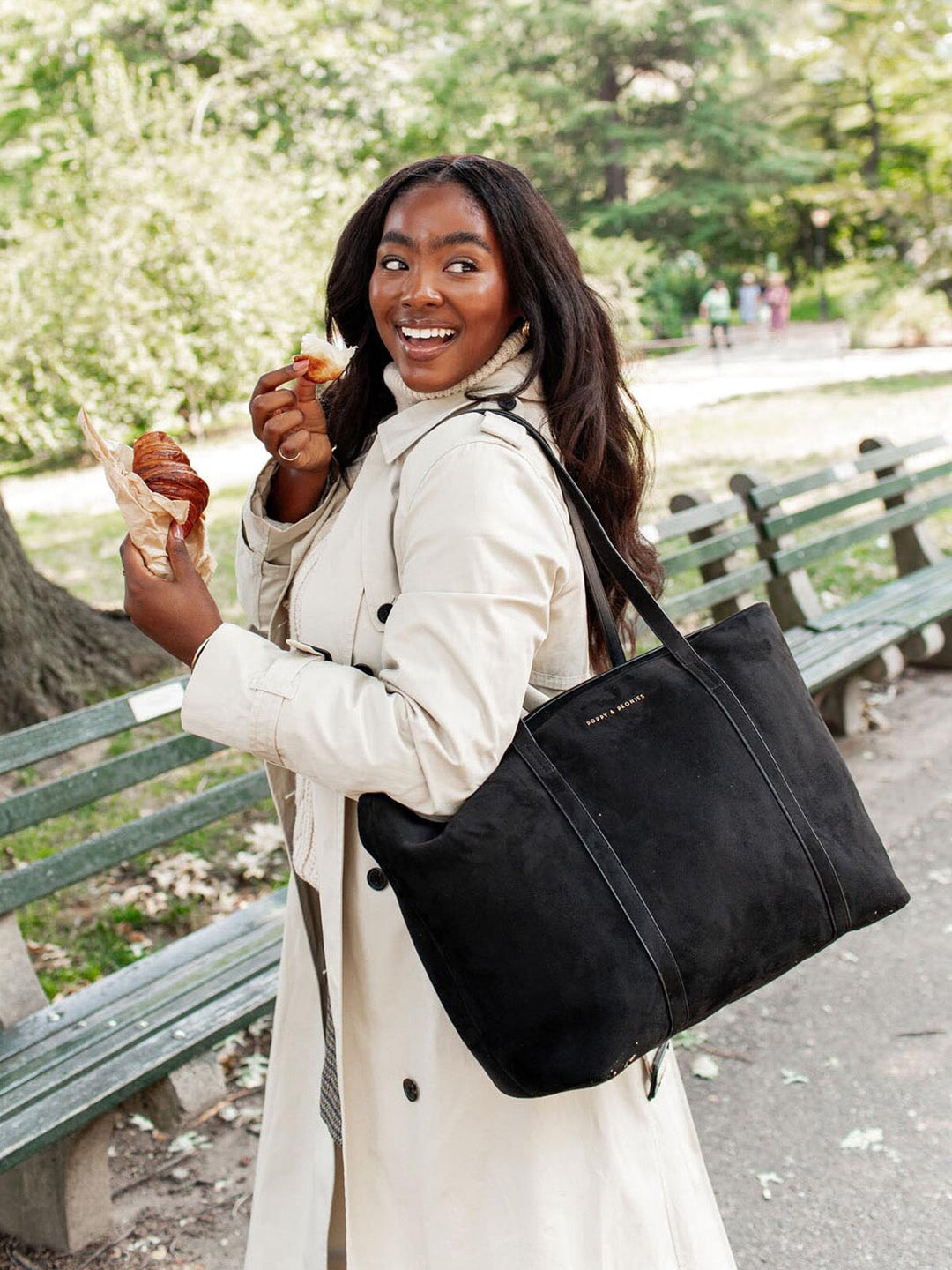 Woman in a park holding a black vegan suede tote bag and eating, with trees and benches in the background