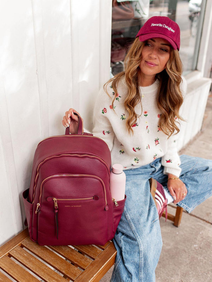 Woman holding a berry coloured backpack wearing a white sweatshirt with floral patterns and a red cap.