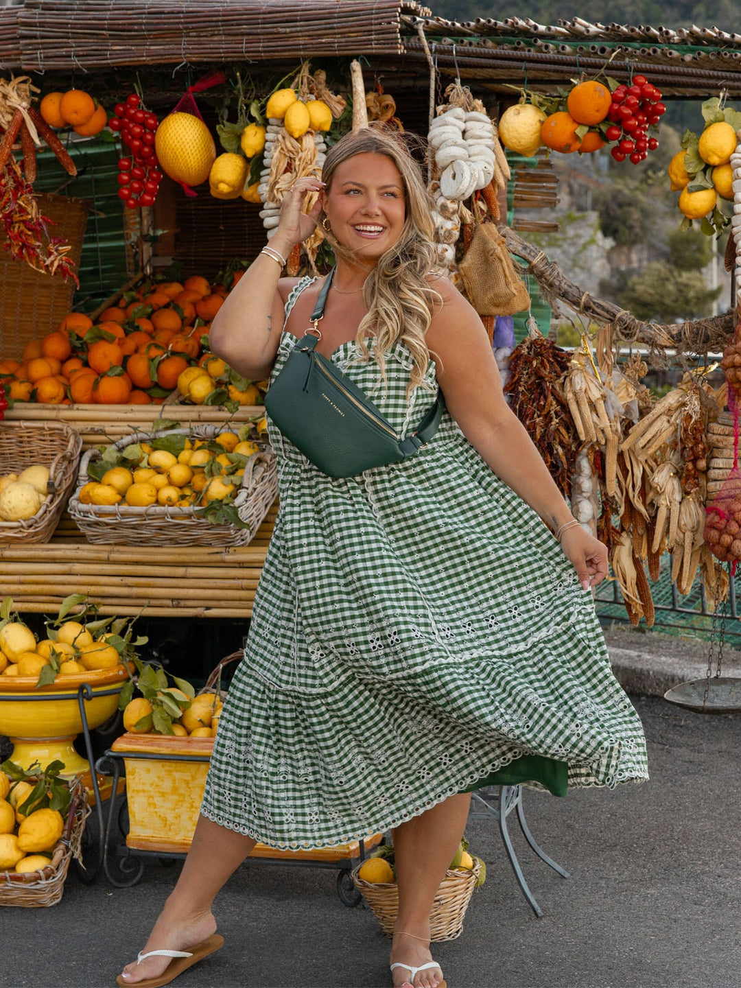 Woman in a green checkered dress and green crossbody bag standing in front of a fruit stand with lemons and other produce.