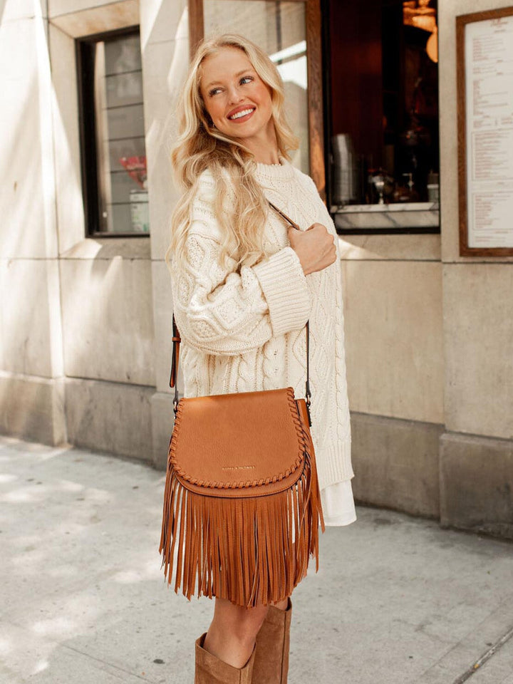 Woman wearing a white sweater and brown fringe bag standing in front of a building.