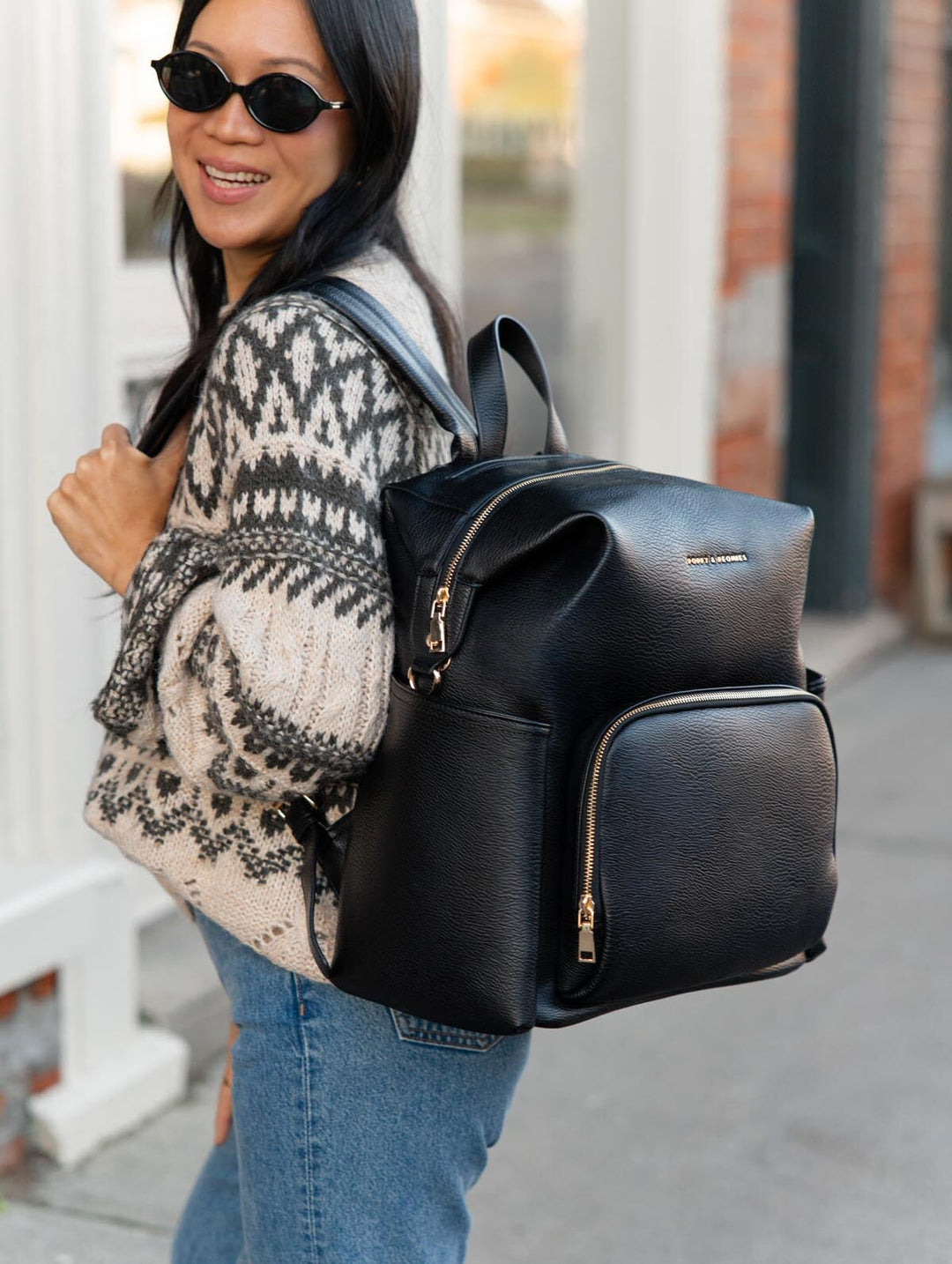 Poppy & Peonies The Cabin Backpack in Black worn over both shoulders by a smiling woman in sunglasses, showcasing the padded shoulder straps and gold-tone zipper details.