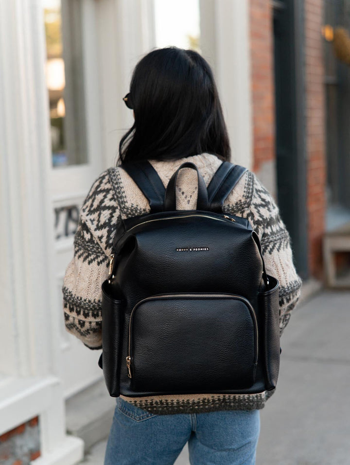 Poppy & Peonies The Cabin Backpack in Black worn on both shoulders by a woman in a patterned knit sweater and blue jeans, shown from behind on a city sidewalk.