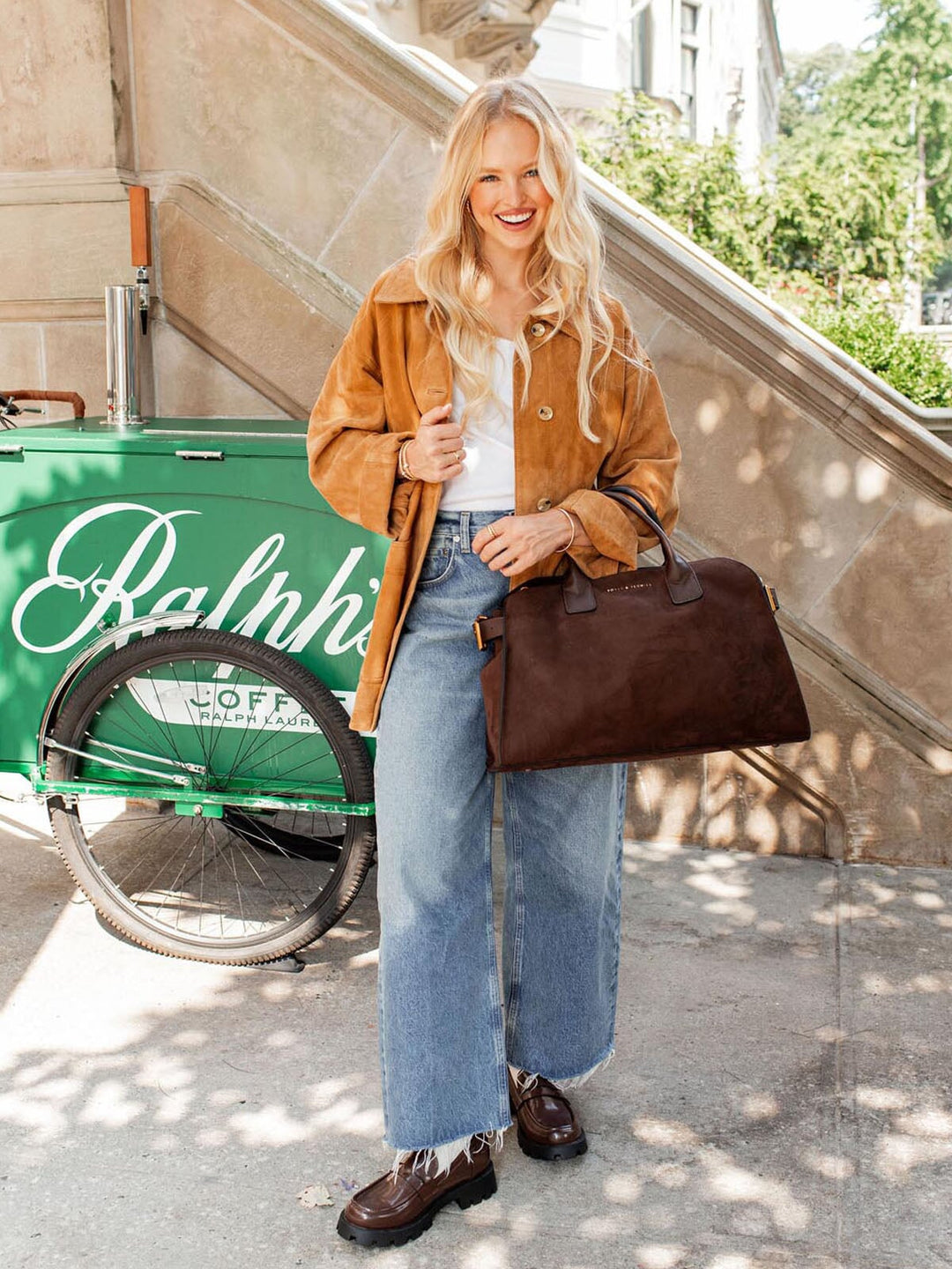 Woman in a brown jacket and blue jeans holding a brown vegan suede bag, standing next to a green coffee bike.