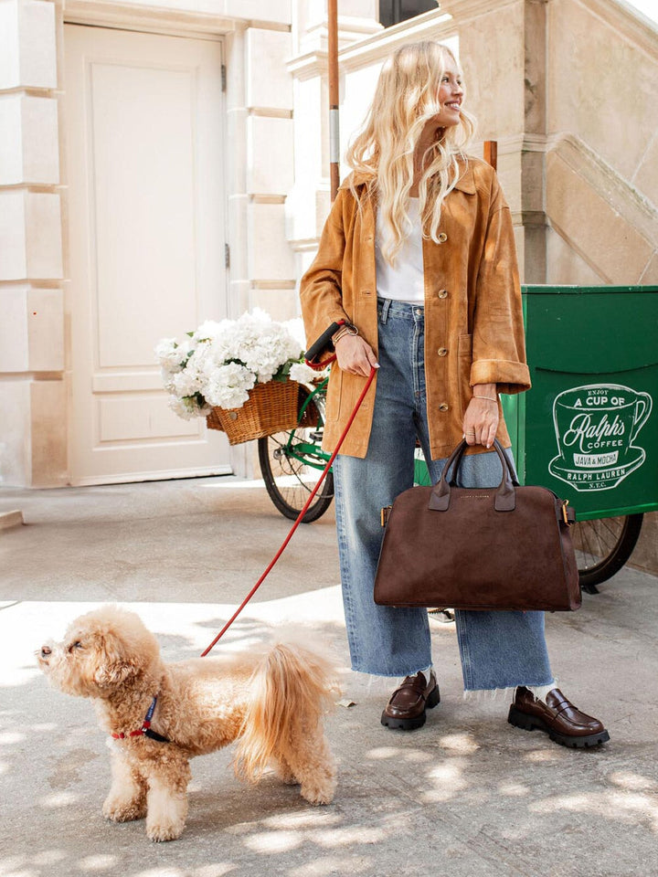 Woman walking a dog on a leash with a brown vegan suede bag, standing next to a green coffee cart.
