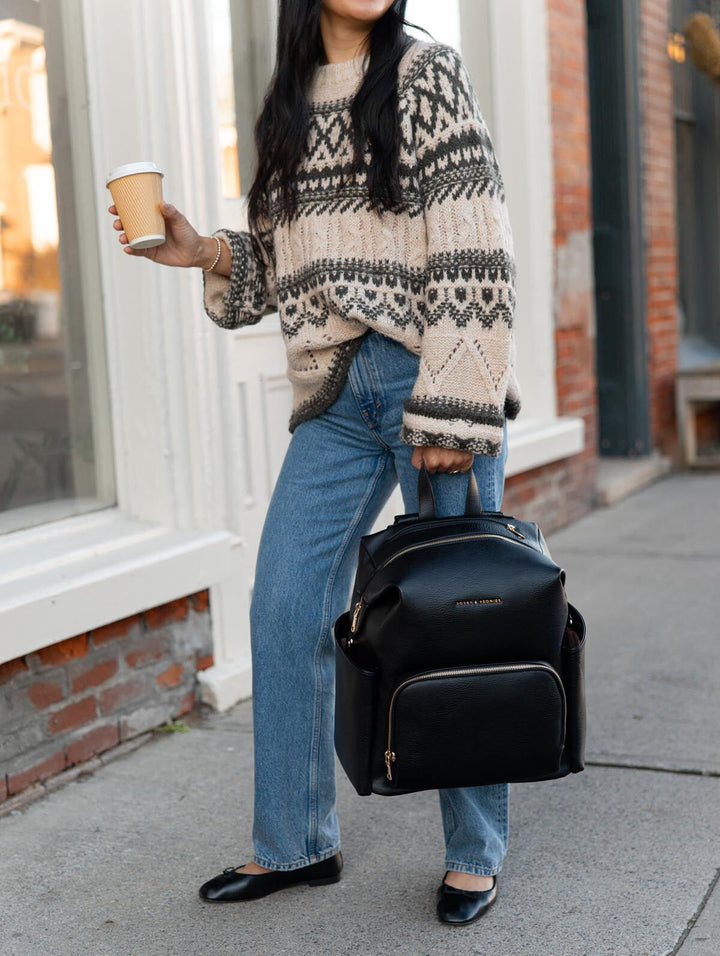 Poppy & Peonies The Cabin Backpack in Black carried by its top handle by a woman in a patterned sweater and jeans, highlighting the large front zippered pocket and structured silhouette.