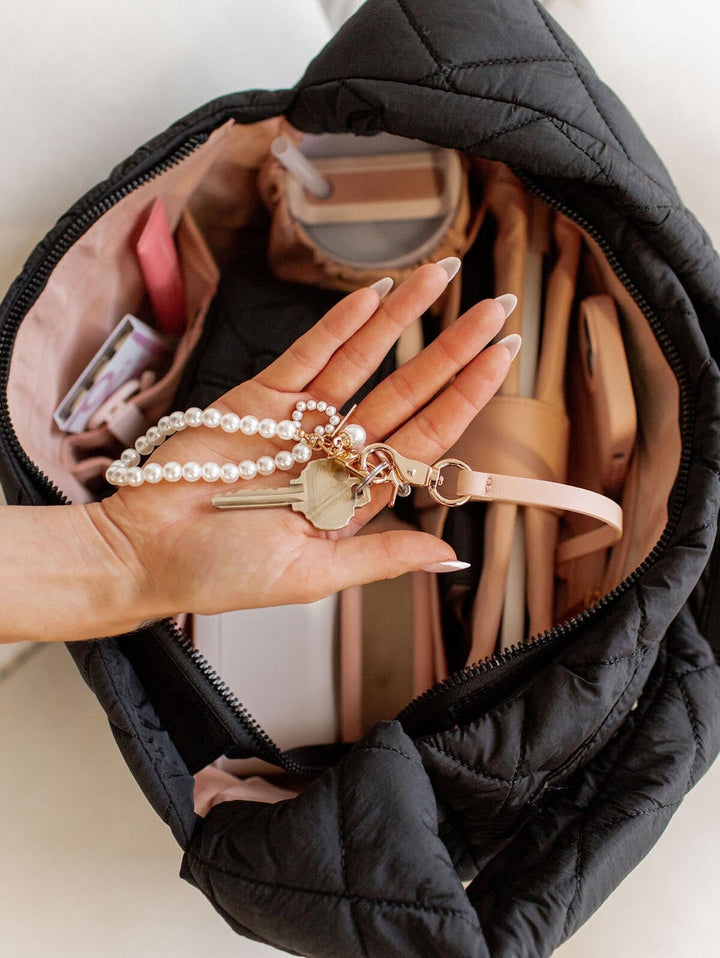 Hand holding keys with a pearl charm in front of an open black quilted handbag.