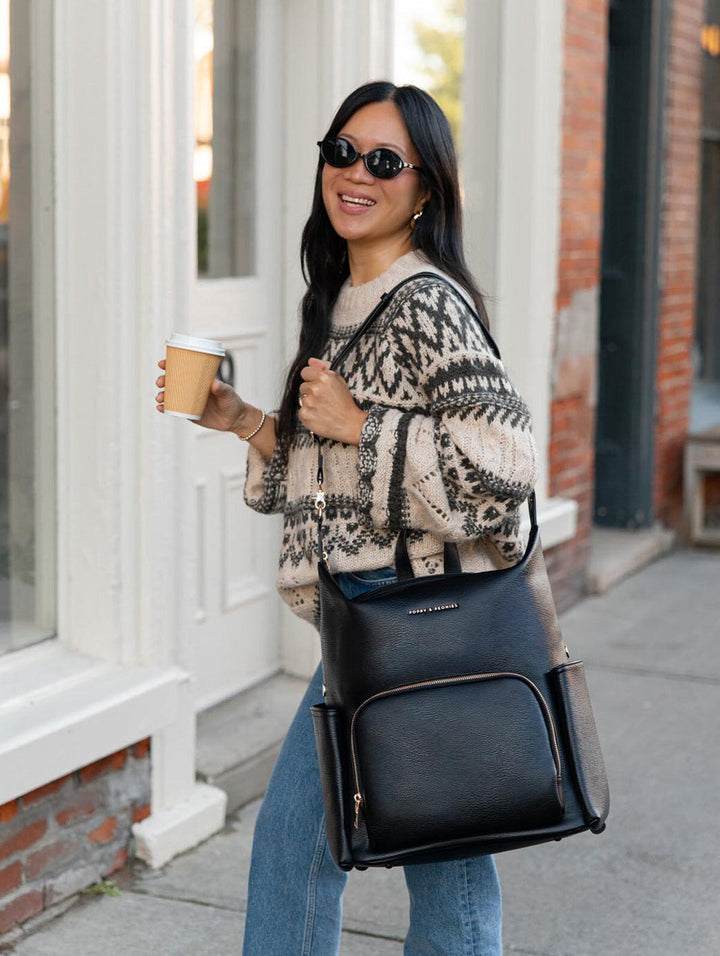 Poppy & Peonies The Cabin Backpack in Black carried over one shoulder by a woman wearing sunglasses, a patterned knit sweater, and blue jeans while holding a takeaway coffee.