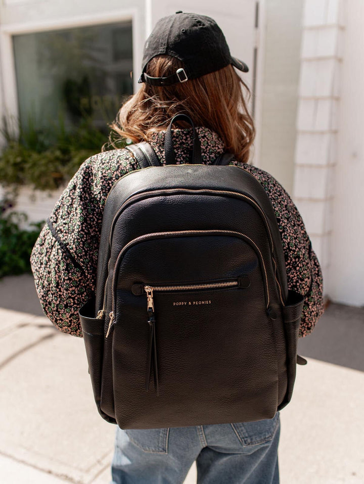Person wearing a black backpack with Poppy & Peonies branding, standing outdoors.