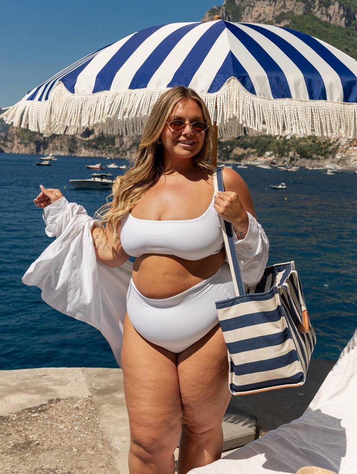 Woman in a white bikini and striped tote bag under a blue and white striped umbrella by the sea.