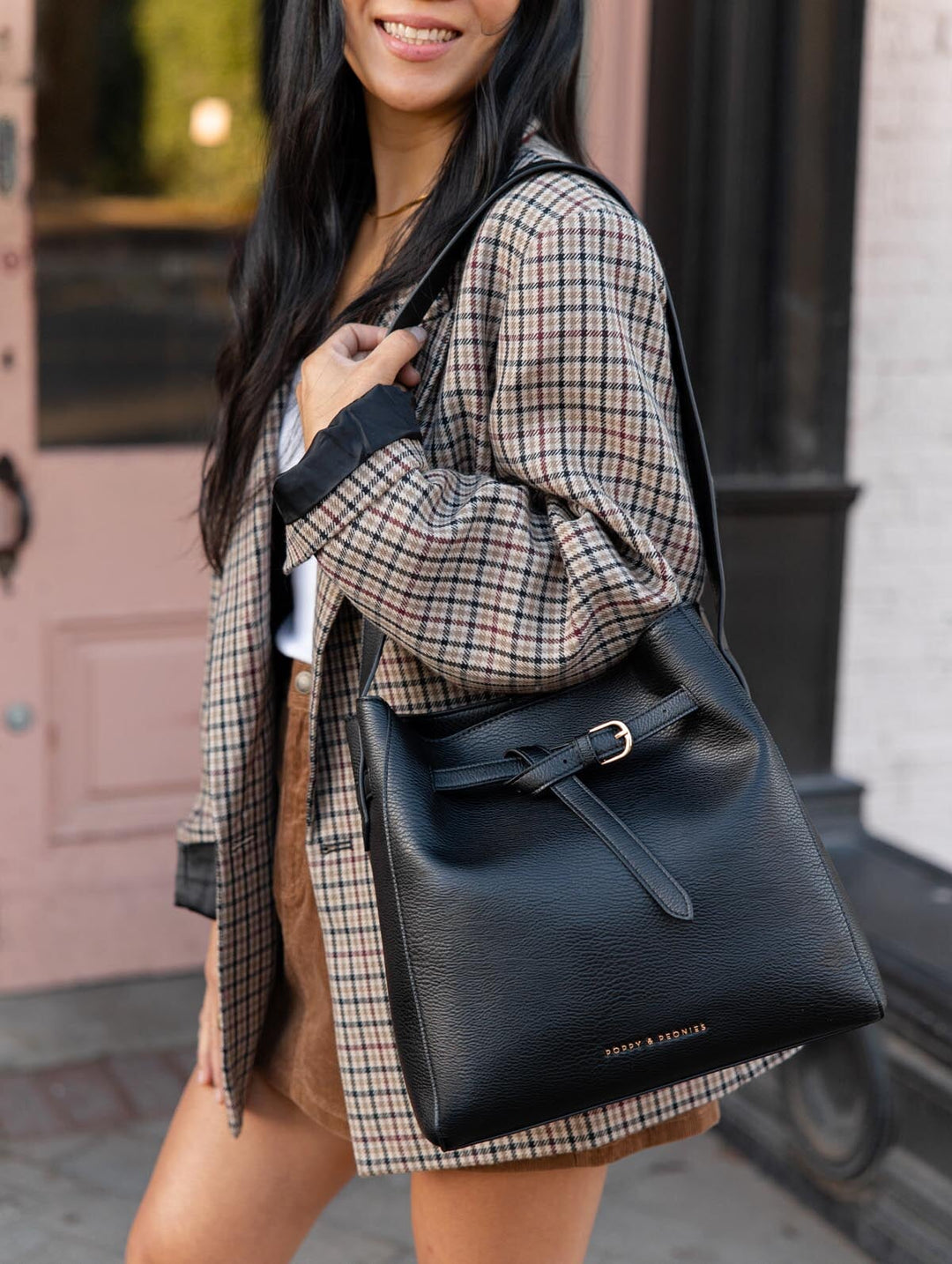 Poppy & Peonies The Knot Shoulder Bag in Black carried over the shoulder by a woman in a plaid blazer, showcasing its spacious silhouette and decorative front belt.