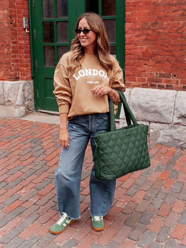 Woman wearing a beige sweatshirt with 'London England' text, blue jeans, and green sneakers, holding a green quilted tote bag on a brick street.