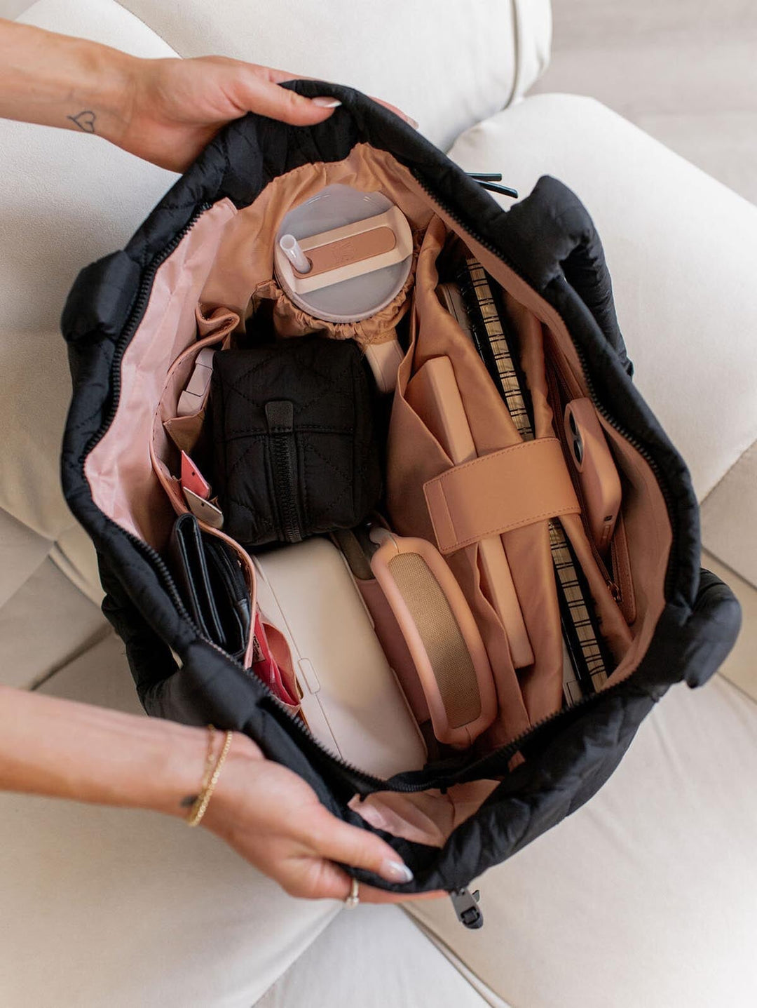 Person opening a black quilted tote bag with brown and beige items inside on a light-coloured floor.